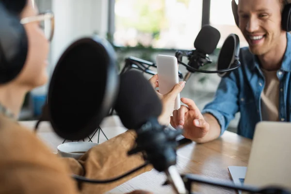 Heureux Hôte Radio Pointant Sur Smartphone Près Flou Asiatique Collègue — Photo