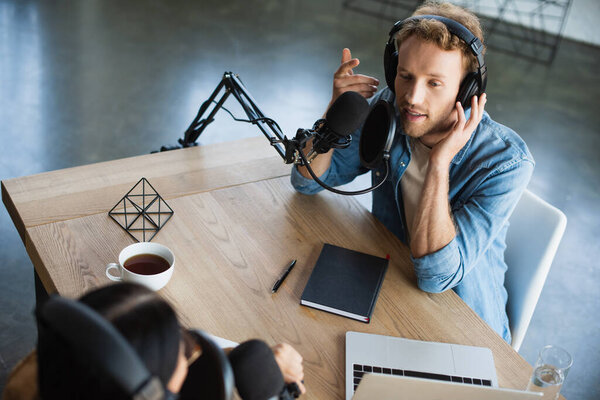 высокий угол обзора радиоведущего, говорящего в микрофоне и жестикулирующего возле ноутбука и коллеги во время подкаста