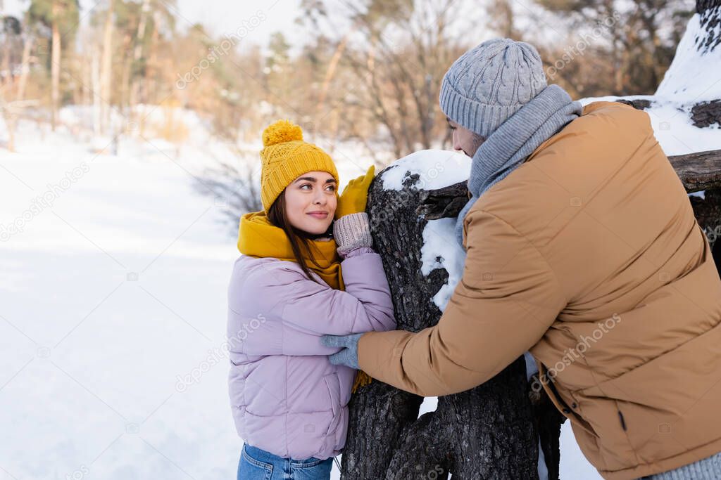 Young woman looking at boyfriend in winter outfit near tree in park 