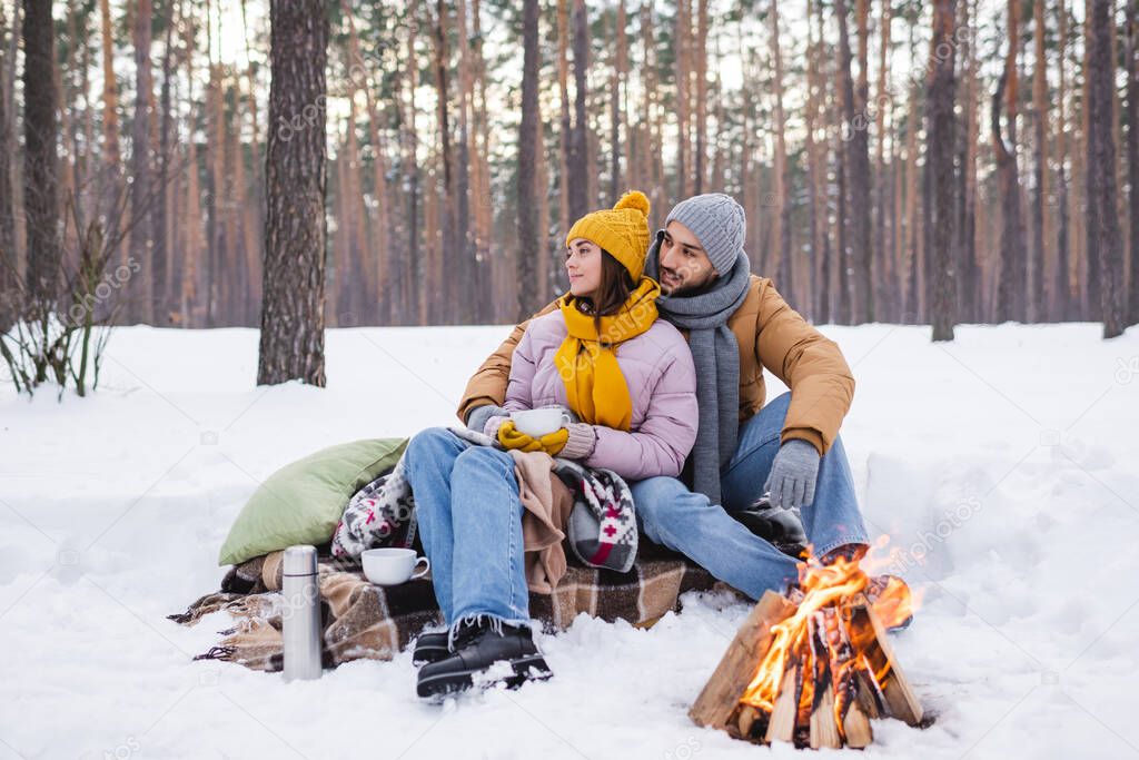 Young couple in winter outfit looking away near cups and bonfire in winter park 
