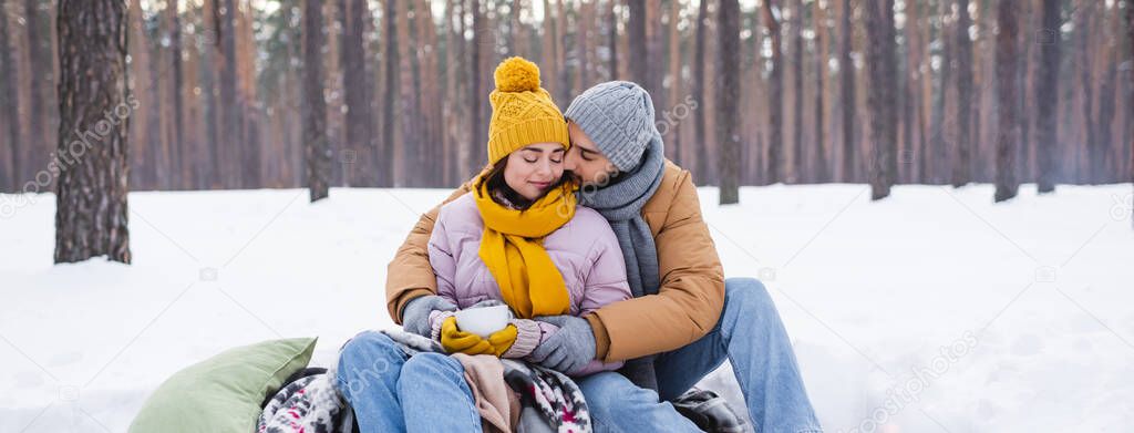 Young man kissing girlfriend in winter outfit with cup in snowy park, banner 