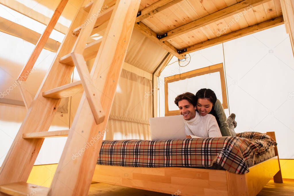 Smiling interracial couple using laptop on bed in glamping house 