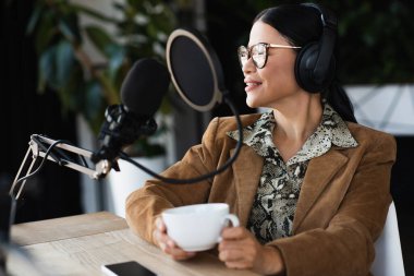 Memnun Asyalı radyo sunucusu gözlüklü ve kulaklıklı podcast sırasında elinde bir fincan kahve tutuyor. 