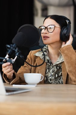 Gözlüklü ve kulaklıklı Asyalı radyo sunucusu podcast sırasında kahve fincanının yanında konuşuyor. 