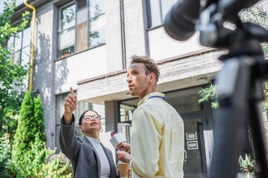 distracted reporter holding microphone near asian businesswoman pointing away near blurred digital camera on tripod  clipart