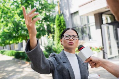 Asyalı iş kadınının yanında elinde kağıt bardakla mikrofon tutan ve röportaj yaparken işaret eden bir muhabir. 