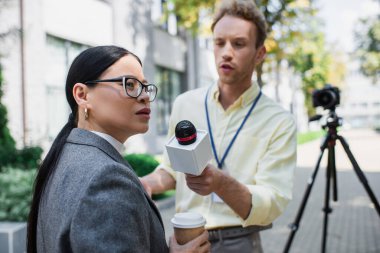 reporter holding microphone near distracted asian businesswoman on street  clipart