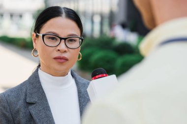asian businesswoman in glasses looking at camera near blurred journalist  clipart