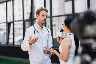 Beyaz önlüklü doktor mikrofonlu Asyalı gazeteciyle röportaj yaparken el kol hareketi yapıyor. 