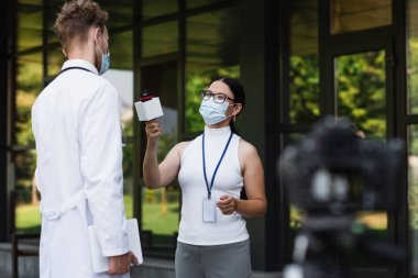 Tıbbi maskeli Asyalı muhabir mikrofonu beyaz önlüklü doktorun yanında tutuyor. 