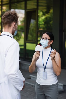 Tıbbi maskeli Asyalı muhabir mikrofonla konuşuyor. Beyaz önlüklü, dijital tabletli doktorun yanında. 