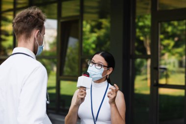 Tıbbi maskeli Asyalı muhabir, kliniğin yanındaki beyaz önlüklü doktorun yanında mikrofonla konuşuyor.