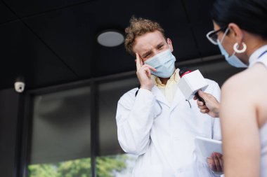 Tıbbi maskeli Asyalı muhabir mikrofon tutuyor. Yanında da dijital tabletli rahatsız edici beyaz önlüklü doktor röportaj veriyor. 