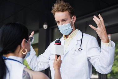 Bulanık Asyalı muhabirin sorgusu sırasında tıbbi maskeli doktor el kol hareketi yapıyor.