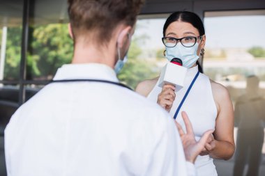 Gözlüklü ve tıbbi maskeli Asyalı muhabir mikrofonda konuşuyor. Beyaz önlüklü bulanık doktor yanında.