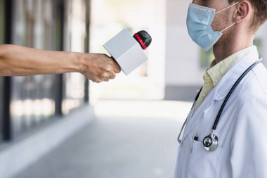 Tıbbi maskeli bir doktorun yanında mikrofon tutan muhabir görüntüsü