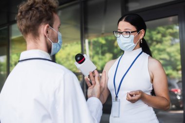 Esmer Asyalı gözlüklü ve tıbbi maskeli muhabir doktorun yanında mikrofon tutuyor. 