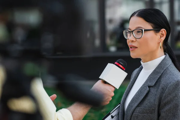 reporter holding microphone and taking interview of asian businesswoman in glasses outside