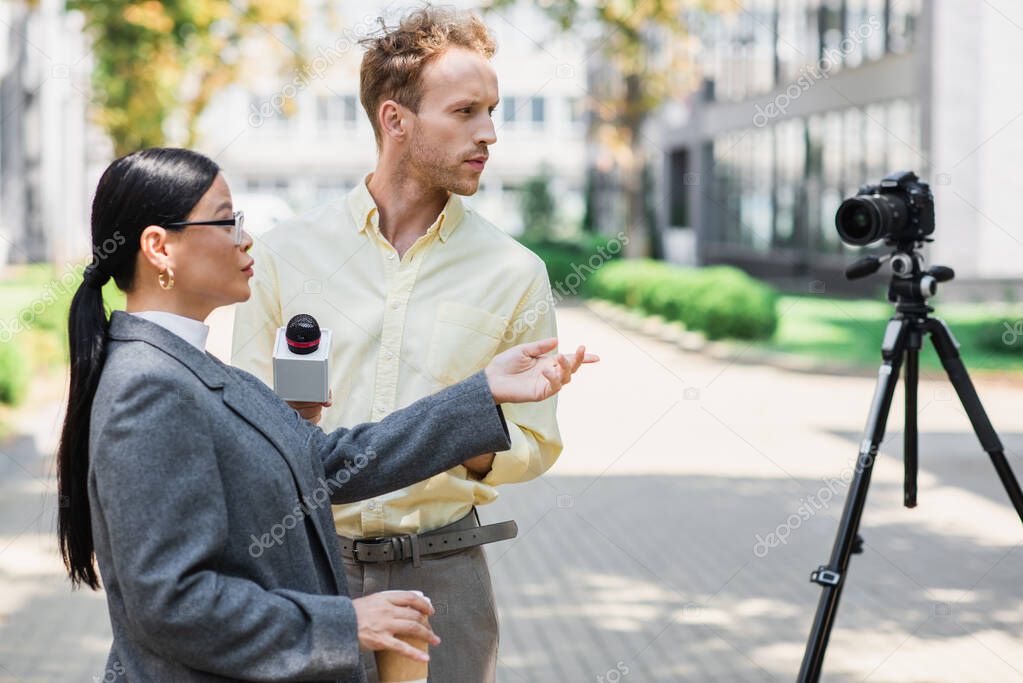 reporter holding microphone near asian businesswoman pointing at digital camera on tripod 