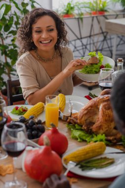 Happy african american mother holding salad near blurred father and traditional thanksgiving dinner  clipart