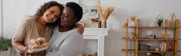 Χαμογελώντας Αφροαμερικανή Γυναίκα Κρατώντας Ψωμάκια Και Αγκαλιάζοντας Μητέρα Στο Σπίτι — Φωτογραφία Αρχείου