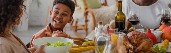 笑着的非洲裔美国男孩看着妈妈在感恩节大餐时拿着沙拉 打着横幅 — 图库照片