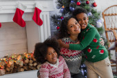 happy african american woman with kids hugging near blurred christmas tree and decorated fireplace