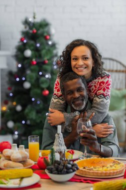 Mutlu bir Afro-Amerikan kadını Noel yemeğinde elinde bir bardak kırmızı şarapla babamı kucaklıyor.