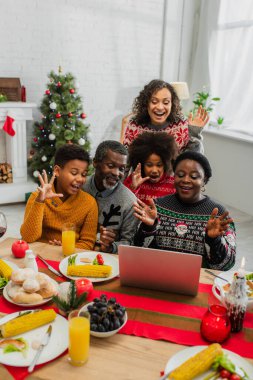 Heyecanlı Afro-Amerikan ailesi Noel yemeği sırasında video görüşmesi yaparken el sallıyor.