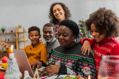Orta yaşlı Afrikalı Amerikalı bir kadın Noel yemeği sırasında laptopunda neşeli bir ailenin yanında daktilo yazıyor.