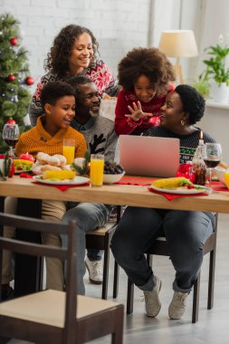 Afro-Amerikalı kız, Noel yemeğinde dizüstü bilgisayar ve mutlu bir aile ile el sallıyor.