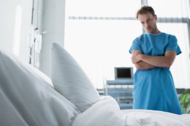 hospital bed with white bedding near sad man in patient gown standing with crossed arms on blurred background  clipart