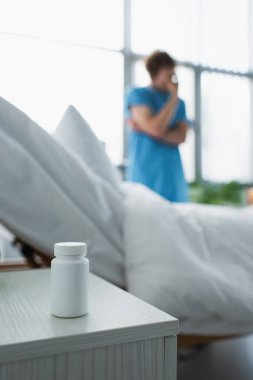 bottle with medication on bedside table near blurred patient in hospital  clipart