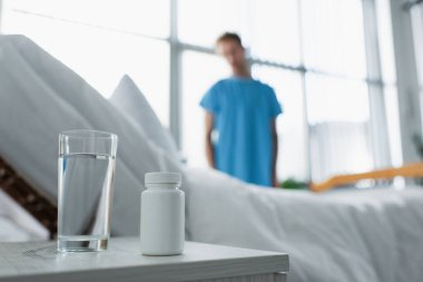 bottle with medication and glass of water on bedside table near blurred patient in hospital  clipart