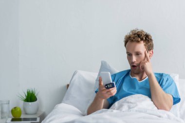 Hastanede akıllı telefon kullanan parmak oksimetresi olan şaşırmış bir hasta. 