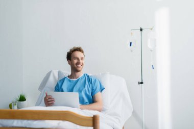 cheerful freelancer in patient gown using laptop in hospital bed  clipart