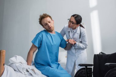 Gözlüklü doktor hastaya tekerlekli sandalyenin yanındaki yataktan kalkması için yardım ediyor. 