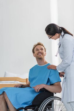 Beyaz önlüklü doktor tekerlekli sandalyedeki neşeli engelli hastanın yanında. 