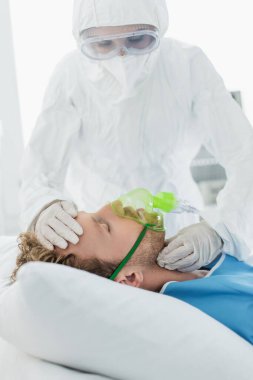 Tehlikeli madde giysisi ve gözlüklü doktor hastanedeki oksijen maskeli hastayı muayene ediyor. 