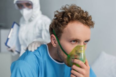 blurred doctor in hazmat suit calming sick patient breathing in oxygen mask  clipart