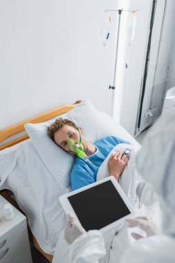 Yüksek açılı oksijen maskeli hasta tehlikeli madde giysisi ve dijital tabletli lateks eldivenli doktora bakıyor. 