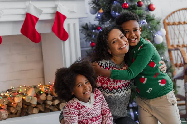 子供を持つ幸せなアフリカ系アメリカ人女性ぼやけたクリスマスツリーと装飾された暖炉の近くに抱きつく — ストック写真