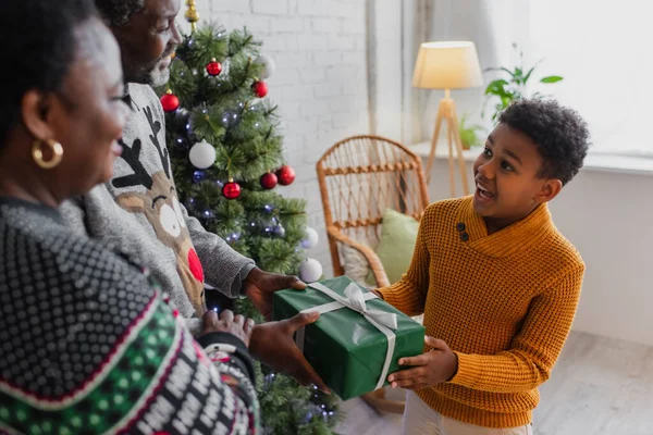 Amazed African American Boy Taking Christmas Gift Blurred Grandparents — Stock Photo, Image