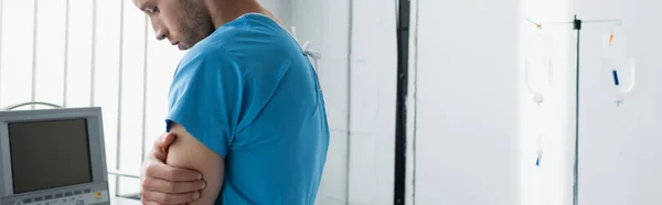 病院で腕を組んで立っている患者のガウンを着た悲しい男バナー — ストック写真
