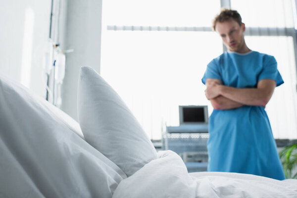 hospital bed with white bedding near sad man in patient gown standing with crossed arms on blurred background 