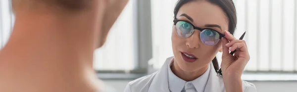 Brunette Doctor Pen Adjusting Glasses While Looking Blurred Patient Hospital — Stock Photo, Image