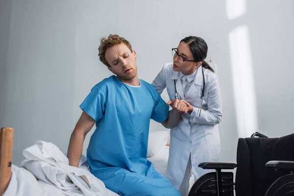 врач в очках помогает пациенту встать с больничной койки возле инвалидного кресла 