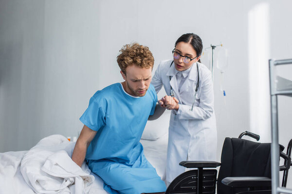 doctor in glasses helping curly patient get up from hospital bed near wheelchair 