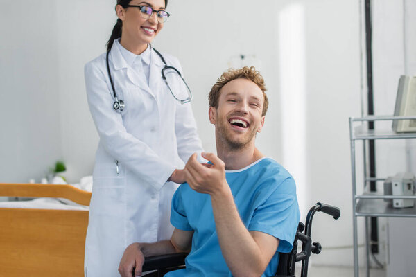 doctor in white coat and glasses smiling near cheerful disabled patient in wheelchair 