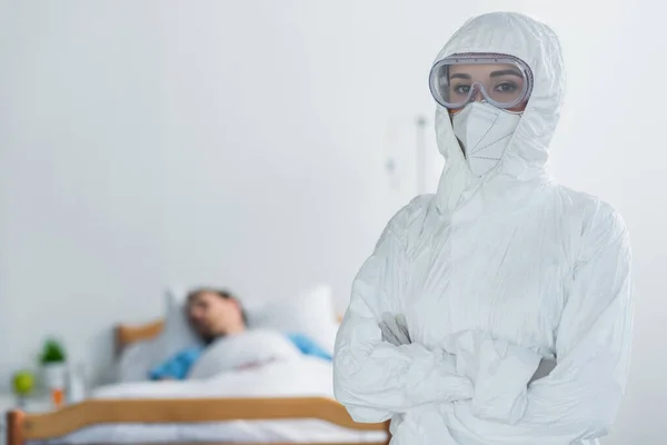 doctor in personal protective equipment and goggles standing with crossed arms near blurred patient in hospital bed